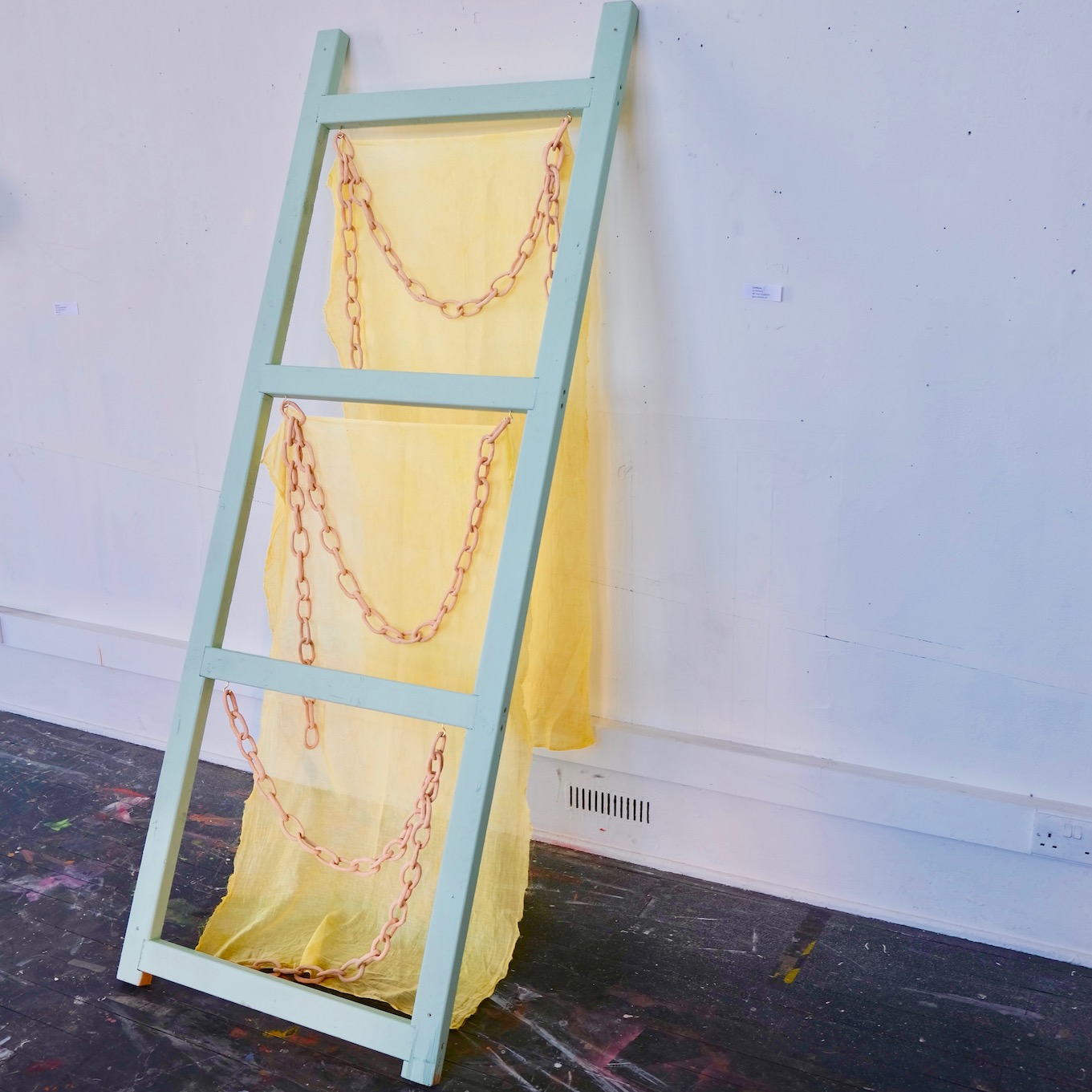Image of sculpture leaning against the wall. It resembles a ladder with fabric hanging from each rung and ceramic chains draped.