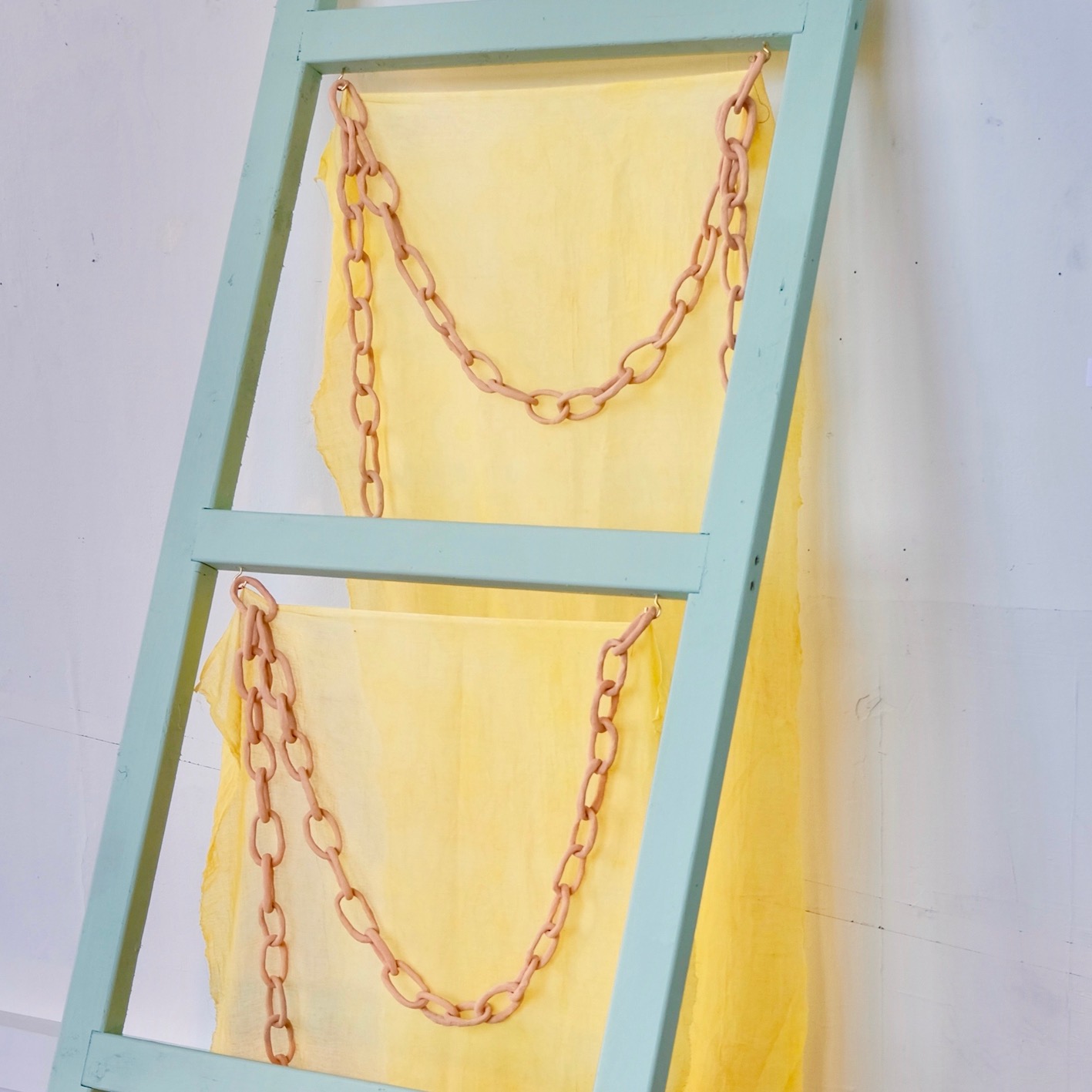 Image of sculpture leaning against the wall. It resembles a ladder with fabric hanging from each rung and ceramic chains draped.