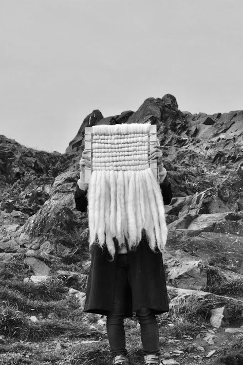 Photograph of weave made from Shetland sheep's wool and hands-on, hand-dyed thread on a wooden frame. The weave is held up by a model whose face is covered by the weave and they are set against the background of the crags.
