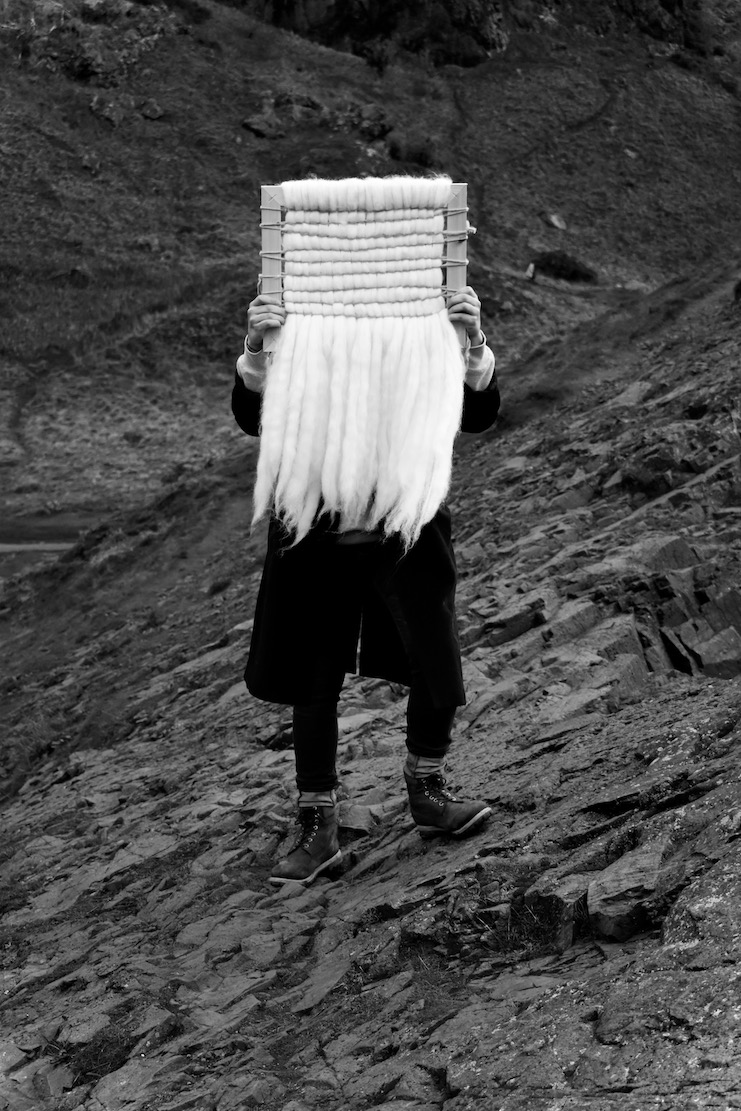 Photograph of weave made from Shetland sheep's wool and hands-on, hand-dyed thread on a wooden frame. The weave is held up by a model whose face is covered by the weave and they are set against the background of the crags.