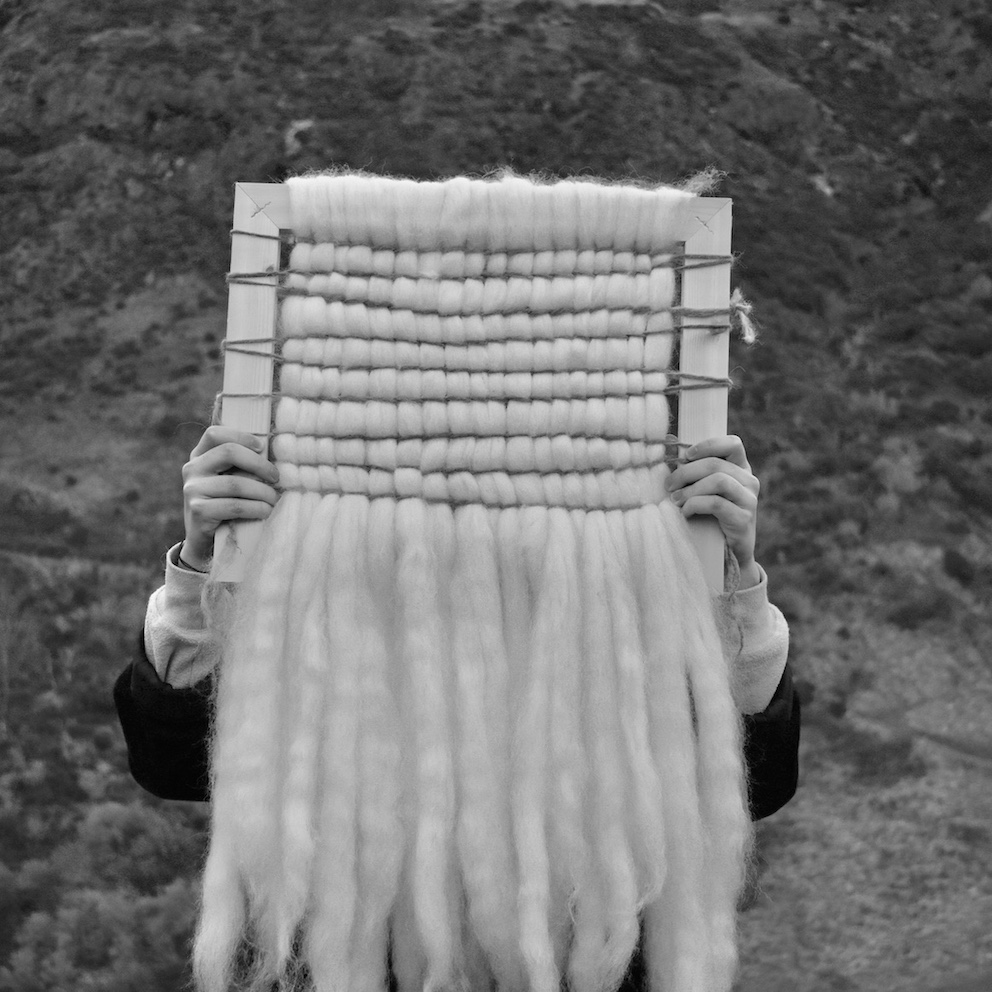 Photograph of weave made from Shetland sheep's wool and hands-on, hand-dyed thread on a wooden frame. The weave is held up by a model whose face is covered by the weave and they are set against the background of the crags.