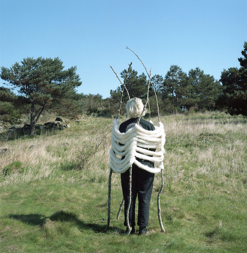 Image of sculpture in the landscape. Branches coming out of the ground with Welsh sheep's wool woven around. Inside the sculpture stands a person, facing away from the camera.