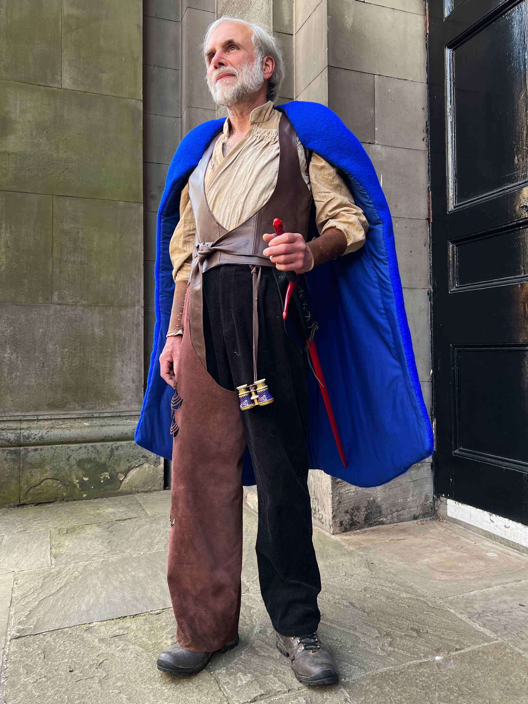 The model wears a light brown shirt that crosses over the body. It is partially covered by a leather waistcoat and on top is a blue cape. 